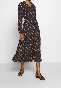 Musta midi mekko kukkakuviolla, jossa on röyhelöhelma, pitkät hihat ja vyötäröä korostava muotoilu, valmistettu kevyestä kankaasta.