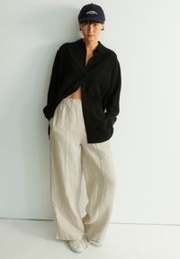 Schwarzes kurzärmliges Hemd, hellbeige weite Leinenhose und weiße Slipper. Eine marineblaue Kappe mit Schriftzug sitzt auf dem Kopf.