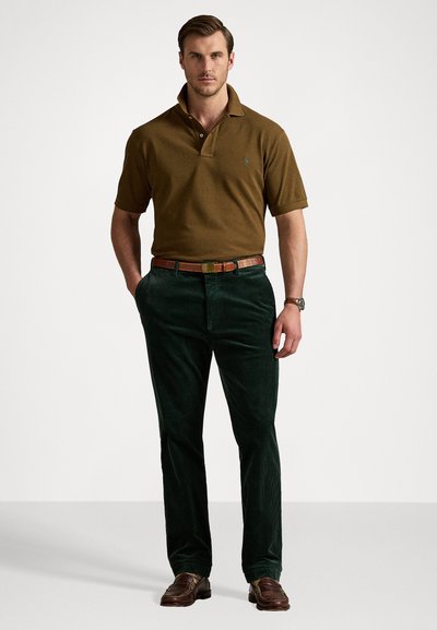 Camisa polo castanha, calças de corduroy verdes escuras, cinto de pele castanho e sapatos castanhos. A polo tem um colarinho e um emblema bordado.