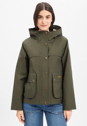 Vrouw draagt een olijfgroene capuchonjas met drukknopen en klepzakken aan de voorkant, gecombineerd met blauwe spijkerbroek, staand tegen een witte achtergrond.