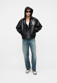 Joven con chaqueta de cuero negra, sudadera con capucha negra, gafas de sol, vaqueros azules y zapatillas blancas, de pie con las manos en los bolsillos.