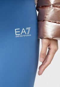 Kék, rugalmas anyagból készült leggings fehér EA7 Emporio Armani logóval. A közelkép egy kézre fókuszál, amely a sima, testhezálló felületen nyugszik.