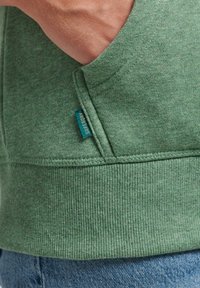 Maglione verde realizzato in tessuto testurizzato con orlo a coste, contrassegnato da un'etichetta con logo visibile in blu. Una mano oscura parzialmente il dettaglio della tasca.