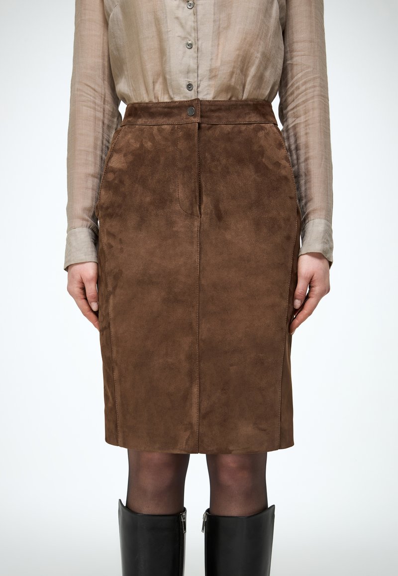 Jupe en daim marron longueur genoux portée avec un chemisier beige transparent à boutons et des bottes noires jusqu'aux genoux, vue de la taille jusqu'aux jambes.