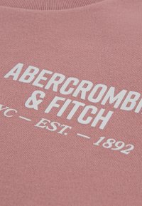 Ροζ πουλόβερ με υφή πλεκτή, με έντονη λευκή επιγραφή «ABERCROMBIE & FITCH NYC - EST. 1892» σε περίοπτη θέση.