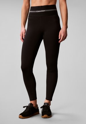 Persoon die zwarte Calvin Klein leggings en zwarte sneakers met bruine zolen draagt, staand tegen een effen lichte achtergrond.