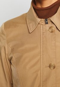 Beige nappatakki, jossa on kaulus ja kaksi suurta nappia. Sileä, hieman kiiltävä kangas; varustettu vetoketjulla kauluksen alla.