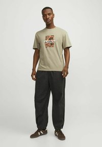 Kakio spalvos trumpas rankovė t-shirt su spalvingu grafiniu spaudiniu ir tekstu priekyje, derinama su laisvai priglundančiomis juodomis kelnėmis ir rudos spalvos sportbačiais.