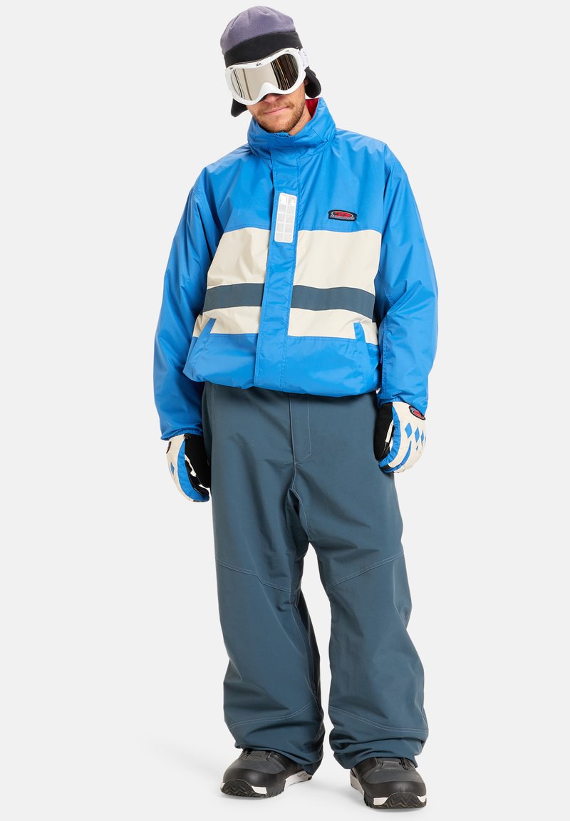 Μπλε σακάκι σκι με λευκές και ναυτικές ρίγες, συνδυασμένο με χαλαρούς μπλε παντελόνε�ς. Εξοπλισμένο με λευκά γυαλιά και μαύρα γάντια.