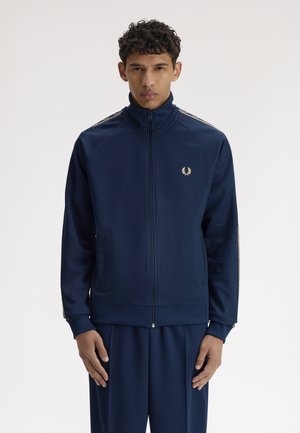 Mann trägt marineblaue Trainingsjacke mit durchgehendem Reißverschluss und passende Hose, steht vor hellem einfarbigem Hintergrund, Arme entspannt an den Seiten.