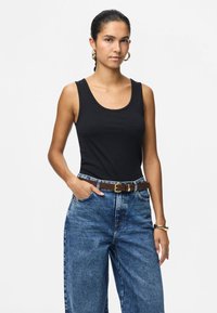 Νεαρή γυναίκα με σκούρα μαλλιά δεμένα σε αλογοουρά, φορώντας χρυσούς κρίκους στα αυτιά, μαύρο τοπ χωρίς μανίκια, μπλε ψηλόμεσα τζιν και καφέ ζώνη, ποζάροντας με το χέρι στην τσέπη.