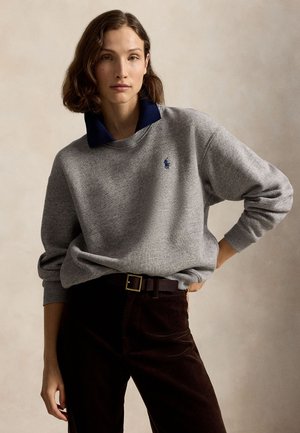 Kvinde med kort brunt hår iført en grå sweatshirt med en mørkeblå krave og mørke bukser, stående med den ene hånd på hoften.
