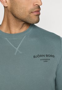 Világoskék pulóver kerek nyakkal, texturált varrással, fekete logóval, amelyen az áll: "BJÖRN BORG STOCKHOLM 1956."