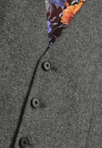 Grå tekstureret blazer med to sorte knapper, der har en farverig blomstermønstret foring, der er synlig ved kraven.