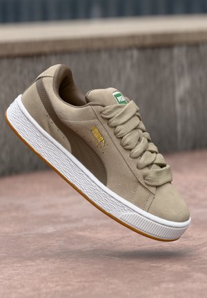 Beige ruskind Puma sneaker med tyk sål og brede snørebånd, i luften over betonjord og sløret stenvæg i baggrunden.