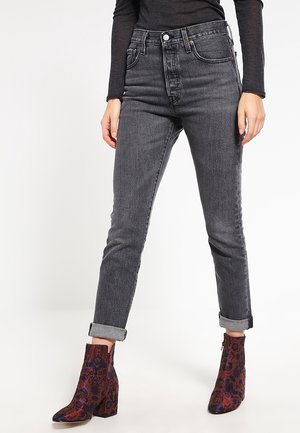 Mujer con vaqueros oscuros remangados, camiseta negra de manga larga y botines estampados con tacones anchos, de pie contra un fondo liso.