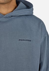 Blå bomulls hettegenser med en myk tekstur, med en liten brodert logo på venstre bryst i en mørkere blå nyanse. Justerbar snøring i hetta.