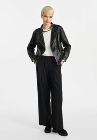 Czarna skórzana kurtka motocyklowa z kontrastowym białym przeszyciem, zestawiona z białą bluzką i czarnymi szerokimi spodniami, noszona z czarnymi butami.