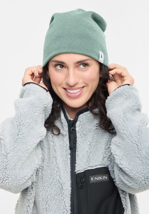Smiling woman adjusting a green beanie, wearing a light gray fleece jacket with black zippered pocket and NIKIN logo.