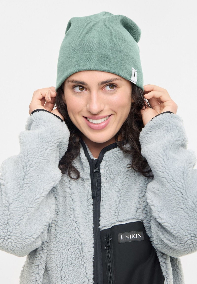 Mujer sonriente ajustándose un gorro verde, vestida con una chaqueta de forro polar gris claro con un bolsillo negro con cremallera y el logo de NIKIN.