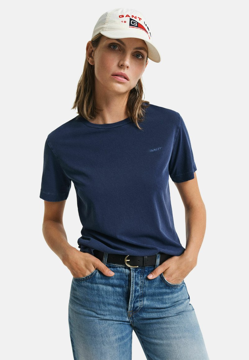 Marineblauw T-shirt met korte mouwen van zachte stof en een ronde halslijn. Het model draagt blauwe jeans en een witte pet met rode en zwarte branding.