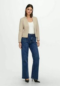 Naine seisab seljas beežist bleiserist, valgest t-särgist, sinistest kõrge vöökohaga teksadest, millel on eespool taskud, ja mustadest teravatest kontsadest, valge tausta ees.