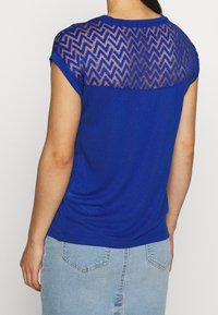 Camiseta azul con un patrón en zigzag texturizado y transparente en el yugo trasero. Manga corta, tejido suave y un ajuste casual. Llevada con denim claro.