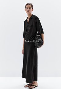 Woman wearing a black collared shirt and long black skirt, carrying a textured black handbag and wearing black sandals, standing against a white background.