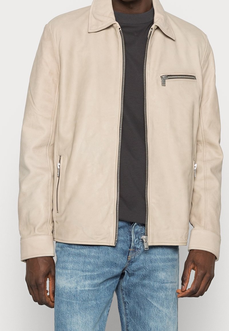 Beige Lederjacke mit kragenförmigem Ausschnitt, Front-Reißverschluss und einer Brusttasche. Verfügt über seitliche Reißverschlusstaschen und eine glatte Textur.