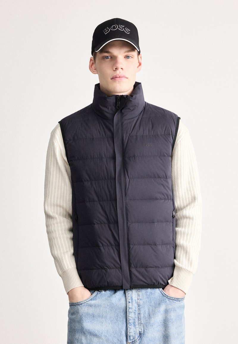 Tumši zila pūķa veste ar augstu apkakli, rāvējslēdzēja aizdari un sānu kabatām. Valkāta pāri gaiši bēša svītrainam džemperim, kombinēta ar zilām džinsa biksēm.