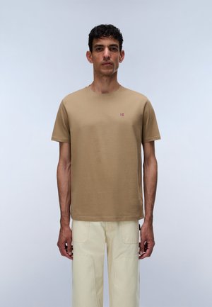 Homme portant un t-shirt à manches courtes couleur sable avec un petit logo drapeau sur la poitrine, associé à un pantalon beige clair sur un fond uni.