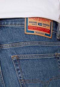 Pantalones vaqueros de mezclilla de color azul, con una etiqueta de cuero marrón en la cintura que muestra detalles de la marca y costuras naranjas contrastantes.