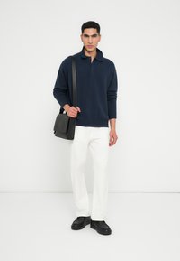 Uomo che indossa un polo a maniche lunghe navy, pantaloni bianchi, scarpe nere e porta una borsa a tracolla nera, in piedi su uno sfondo bianco semplice.