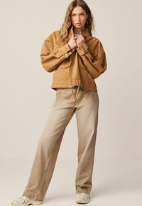 Beige oversize dzseki magas gallérral, patentgombokkal és húzózsinóros aljjal, világosbarna bő nadrággal és fehér sportcipővel párosítva.