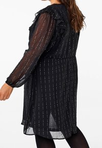 Svart klänning med genomskinliga långärmar, frilldetaljer och vertikala ränder av metallpunkter. Kjolen har en silkeslen textur och en flödande silhuett.