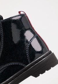 Černé lakované kotníkové boty s lesklým povrchem, kontrastním červeným akcentem na poutku a texturovanou gumovou podrážkou. Obsahují dekorativní prošívání.