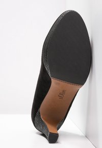 Black suede high heel shoe with a textured leather accent and a smooth black rubber sole, displaying a subtle brand logo on the inner side.