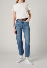 Maglietta bianca in cotone con piccolo logo rosso, jeans azzurri a vita alta, cintura in pelle marrone e scarpe da ginnastica bianche con suole testurizzate.