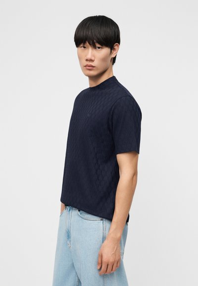 Νεαρός άνδρας με ίσια μαύρα μαλλιά στέκεται πλαγίως φορώντας ένα σκοτεινό κοντομάνικο πουκάμισο με σχέδια και ανοιχτόχρωμο μπλε τζιν, μπροστά σε ένα απλό φόντο.