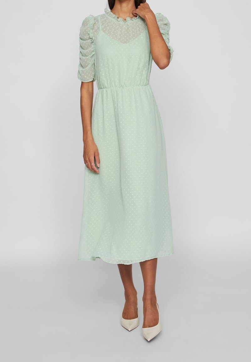 Femme portant une robe midi légère vert clair transparente avec des manches bouffantes, un col à volants, et des talons blancs à bouts pointus, debout devant un fond gris.