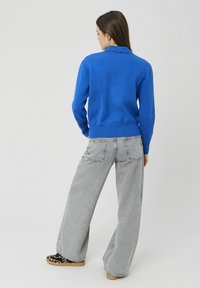 Pull bleu avec un col festonné, associé à un jean gris à jambes larges et des chaussures noires et marron à motifs. Vue de dos.