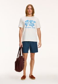 White t-shirt with blue graphic text "HOLIDAY OFTEN," dark blue shorts, brown slip-on shoes, maroon tote bag. Simple design, casual fit.