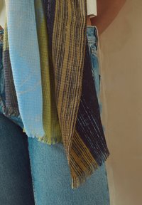 Quatre foulards légers aux tons bleus, verts et bruns suspendus sur un jean en denim bleu clair, porté par une personne, devant un mur beige.