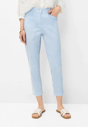 Kvinde iført blå og hvide vertikale stribede korte bukser, hvid bluse med blonder ærmer, guldarmbånd og hvide slip-on sandaler.