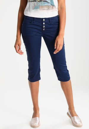Vrouw draagt marineblauwe cropped broek met vier zilverkleurige knopen en lichte instapschoenen, staand tegen een effen achtergrond.