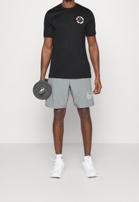 Musta lyhythihainen t-paita, jossa on punainen ja valkoinen logo, yhdistettynä harmaisiin shortsseihin, joissa on huomaamaton Nike-merkki, pitäen mustaa painolautaa.