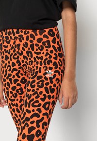 Leggings orange avec un motif léopard noir, dotés d'une taille élastique et d'un logo Adidas blanc sur le bas de la jambe gauche.