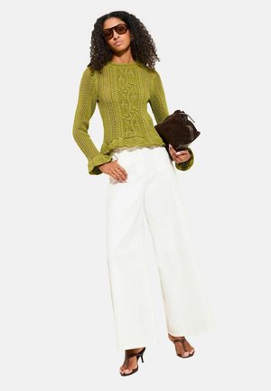 Žena oblečená v zelenom pletenom svetri, bielych širokých nohaviciach, hnedých sandáloch na vysokom podpätku, s okuliarmi proti slnku a držiaca hnedú listovú kabelku.