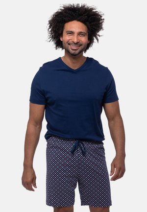 Uomo con capelli ricci che indossa una camicia blu navy a maniche corte e pantaloncini con coulisse a motivo, sorridente su sfondo bianco.