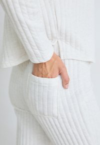 Hand i sidofickan på vita quiltade byxor med matchande quiltad långärmad topp, visad från midjan till höften.